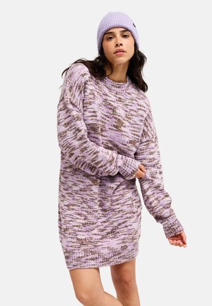 Femme portant une robe-pull tricotée à motif lavande et marron et un bonnet en tricot lavande assorti, posant avec un bras plié et l'autre tendu.