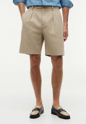 Uomo che indossa pantaloncini beige plissettati con tasche laterali, camicia a maniche lunghe blu arrotolata e mocassini bicolore nero e beige.