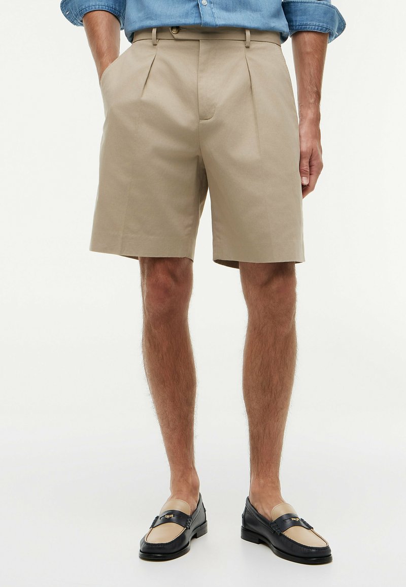 Mann trägt beige plissierte Shorts mit Seitentaschen, blaues hochgekrempeltes Langarmhemd und zweifarbige schwarze und beige Loafers.