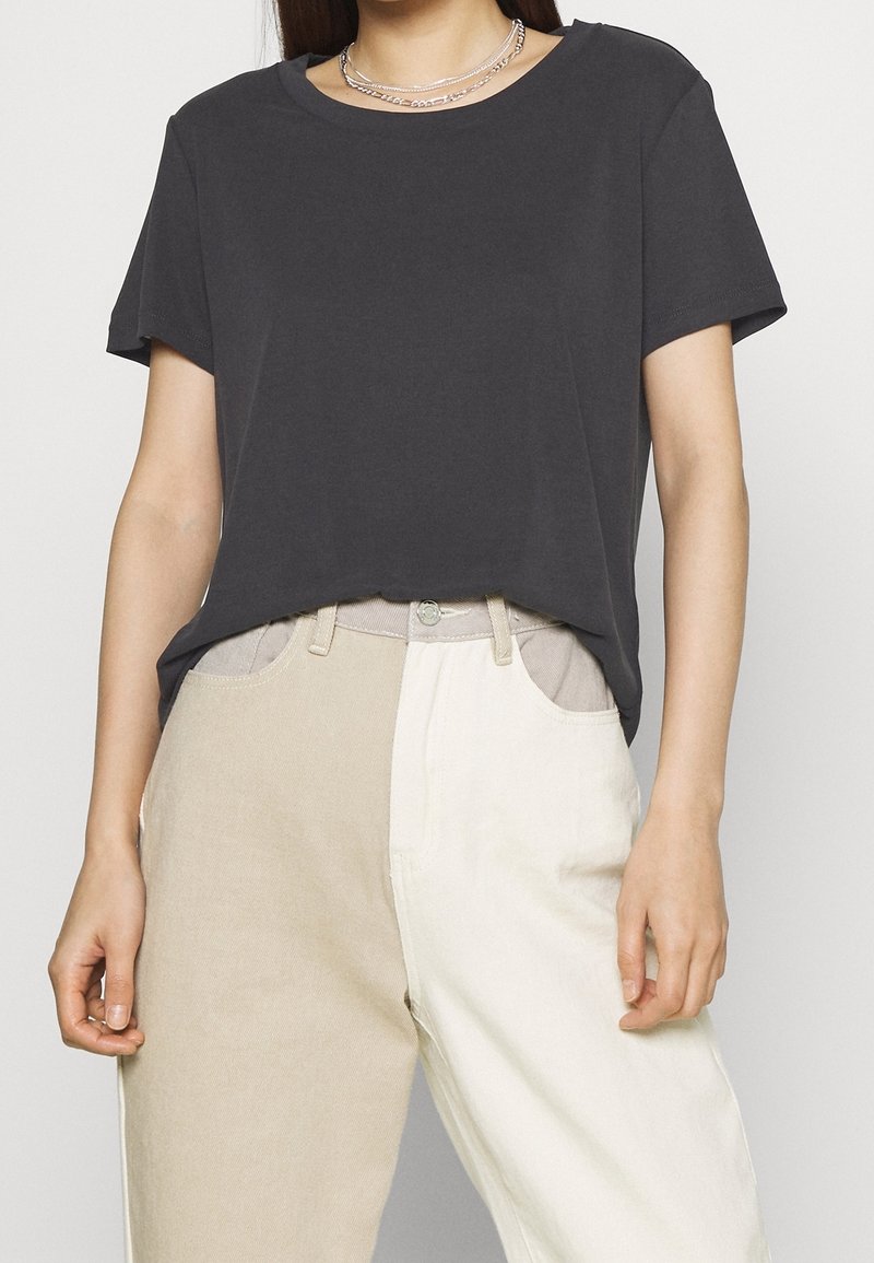 Femme portant un t-shirt noir à manches courtes et un pantalon deux tons beige et blanc, avec des colliers argentés superposés.