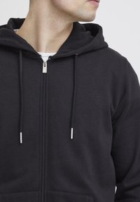Felpa con zip nera realizzata in un tessuto testurizzato, con cordini dotati di aglette in argento e una tasca frontale priva di motivi o loghi visibili.