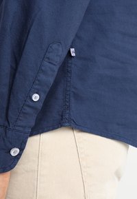 Marineblauw overhemd met lange mouwen en witte knopen, gedragen over beige broek, met zichtbare zijzoom en klein stoffen label.