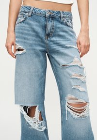 Ανοιχτό μπλε τζιν παντελόνι με ψηλή μέση και πολλές μεγάλες φθορές στους μηρούς και τα γόνατα, φορεμένο από άτομο με τα χέρια ακουμπισμένα δίπλα στο σώμα.