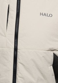 Beżowa kurtka z czarnym zamkiem błyskawicznym, czarnym detalem z siateczki na kieszeni oraz logo "HALO" na klatce piersiowej.