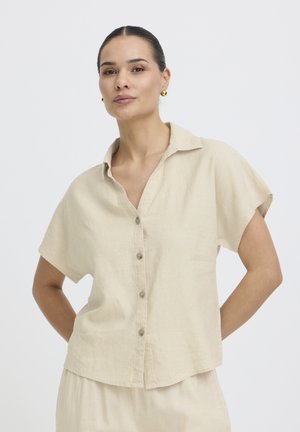 Žena s tmavými vlasmi zviazanými do chvosta, oblečená v béžovej krátkorezej košeli na gombíky a zodpovedajúcich nohaviciach, stojaca s rukami za chrbtom.