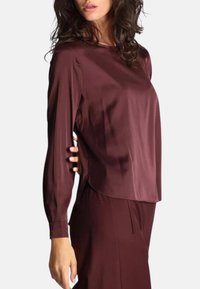 Top lungo in satin bordeaux a maniche lunghe con vestibilità comoda, scollatura rotonda e spacchi laterali, abbinato a pantaloni coordinati sullo sfondo.