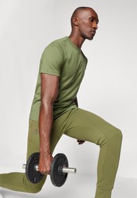 Ελιά πράσινη κοντομάνικη μπλούζα και ταιριαστά αθλητικά παντελόνια, με εφαρμοστή σχεδίαση. Άτομο κρατά μια μαύρη αλτία. Απαλή υφή υφάσματος.