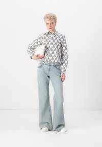 Knappskjorte med svart sirkulært mønster, lyseblå vidbukser og en liten hvit veske. Modellen har på seg hvite sneakers.