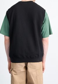 Felpa nera senza maniche con orlo a coste, indossata sopra una camicia verde a maniche corte. Pantaloni cargo color sabbia visibili sotto.