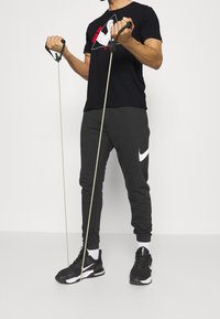 Camiseta negra de Nike con un diseño gráfico, pantalones de chándal grises con un logo blanco y zapatillas deportivas negras que sujetan bandas de resistencia.