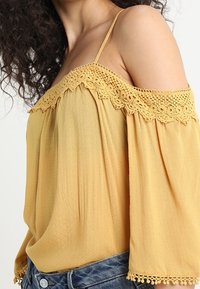 Yellow off-shoulder blouse with lace trim, relaxed fit, and loose sleeves, paired with distressed blue denim shorts. Soft fabric texture.