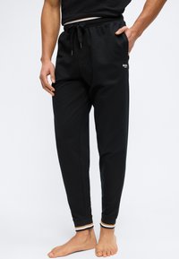 Άνδρας που φορά μαύρο παντελόνι jogger με στενή εφαρμογή, ελαστικά τελειώματα και κορδόνι, με το ένα χέρι στην τσέπη, όρθιος ξυπόλητος σε λευκό φόντο.