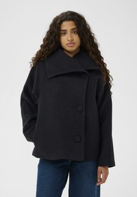 Schwarzer Wollmantel mit weitem Kragen, ausgestattet mit drei großen Knöpfen und lockeren Ärmeln. Der Stoff wirkt weich und strukturiert, mit minimalen Designdetails.