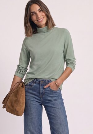 Femme aux cheveux bruns mi-longs portant un col roulé vert clair et un jean bleu, tenant un sac à main en daim marron et souriante.