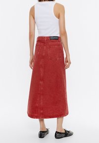 Rode spijkermaxirok met een achtersplit, twee zakken en een hoge tailleband, gecombineerd met zwarte platte schoenen met studs. Witte mouwloze top zichtbaar.