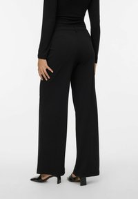 Svarta vida byxor med en slät textur, hög midja och diskret sömnadsdetaljering. Bärs med svarta klackskor.