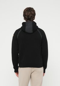 Μαύρο φ ούτερ με λείο υφή, διαθέτοντας αντίθετο μαύρο πάνελ στη κουκούλα και τις ραφές των ώμων. Ριμπ μανσέτες και τελείωμα ολοκληρώνουν το σχέδιο.