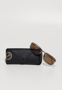Hnedo zafarbené bezrámové slnečné okuliare Ray-Ban so zlatými bočnicami odložené na čiernom koženom puzdre značky Ray-Ban so zlatou nálepkou UV ochrany.