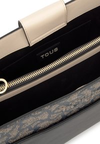 Beżowa i czarna torba wnętrze z widocznym złotym zamkiem, czarnym materiałem podszewkowym oraz metką marki "TOUS" wewnątrz kieszonki.