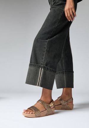 Sandalias metálicas marrones con correas dobles de acabado texturizado y detalle de hebilla lateral, combinadas con jeans oscuros cortos con costuras blancas.