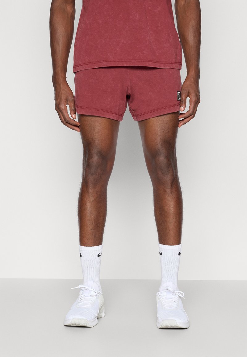 Bordeauxfarbene Shorts mit lockerer Passform, aus weichem Stoff, kombiniert mit weißen Sportsocken, die ein schwarzes Logo aufweisen, und weißen Sneakers.