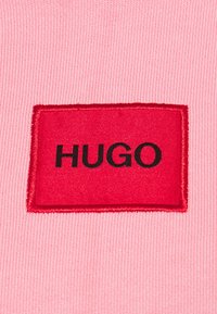 Tissu côtelé rose avec un patch rectangulaire rouge portant le mot « HUGO » en lettres noires grasses centrées.