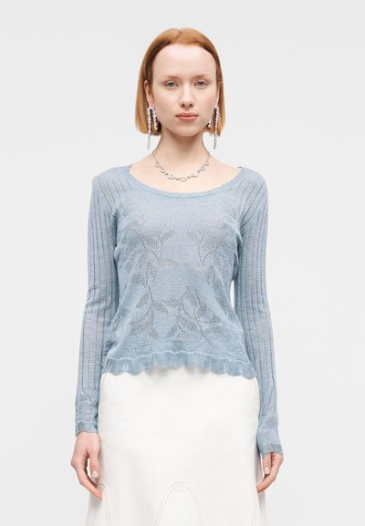 Pull tricoté bleu clair avec motif floral, manches longues, texture côtelée et ourlet festonné, associé à une jupe blanche.