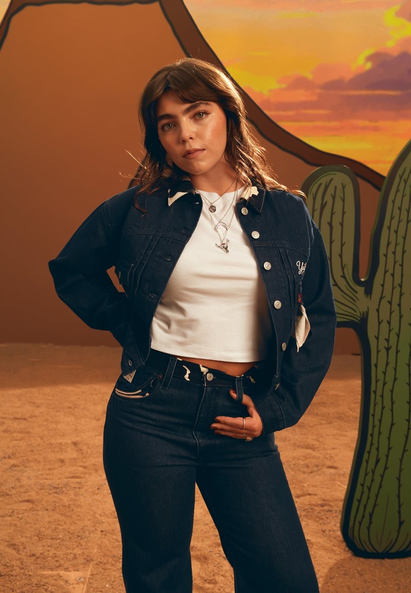 Giacca di jeans con colletto bianco, abbinata a un crop top bianco e jeans scuri con una vita decorata. Sullo sfondo ci sono dei cactus.