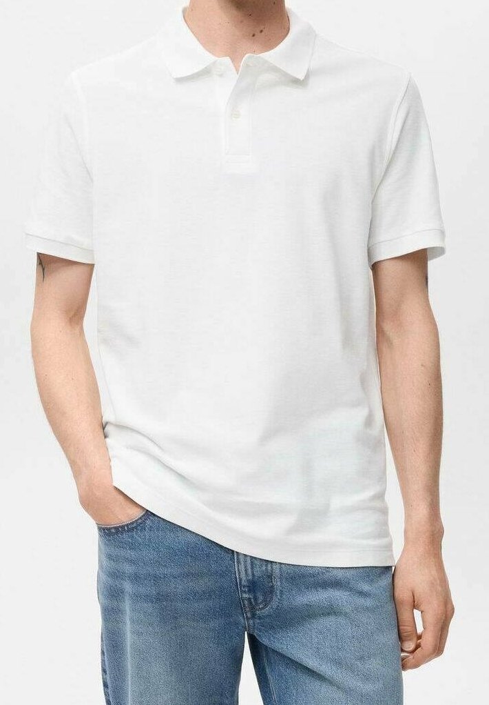 Homme portant un polo blanc uni à manches courtes et un jean bleu, main dans la poche, debout devant un fond clair uni.