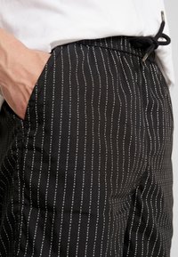 Juodos kelnės su baltu vertikaliu tekstu. Pagamintos iš minkštos medžiagos, turi elastinę liemenį ir reguliuojamą raištį patogumui.