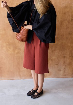 Moteris su blondiniais plaukais, dėvinti juodus pūstus rankovių detalius drabužius, rūdžių spalvos šortus iki kelių ir juodus austa plokščiadugnius batus, laiko rudą austi krepšį kibiro formos.