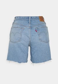 Short en jean bleu clair avec ourlets effilochés, deux poches arrière, un patch de marque en cuir et une petite étiquette rouge sur la poche droite.