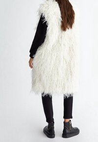 Weste aus Kunstfell in Weiß mit langem, strubbeligem Textur, kombiniert mit schwarzen Hosen und eleganten schwarzen Stiefeletten mit goldenen Akzenten.