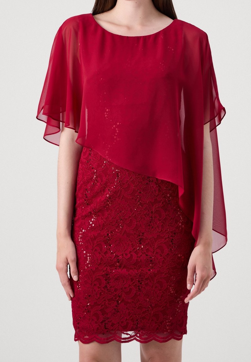 Femme portant une robe rouge en dentelle avec des détails en sequins et un surcroît asymétrique en chiffon transparent avec des manches flottantes.