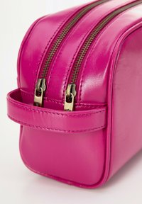 Růžová kožená kosmetická taška se dvěma zlatými zipy, zaoblenou horní částí a bočním madlem. Její hladký povrch a prošití jsou viditelné.