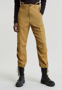 Pantaloni cargo beige con vita alta, tasche laterali e polsini elasticizzati; abbinati a stivali neri con lacci e a un top nero aderente.