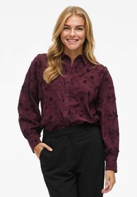 Blouse bordeaux avec broderie florale, design à boutons et manches longues ; associée à un pantalon noir pour un look contrasté.