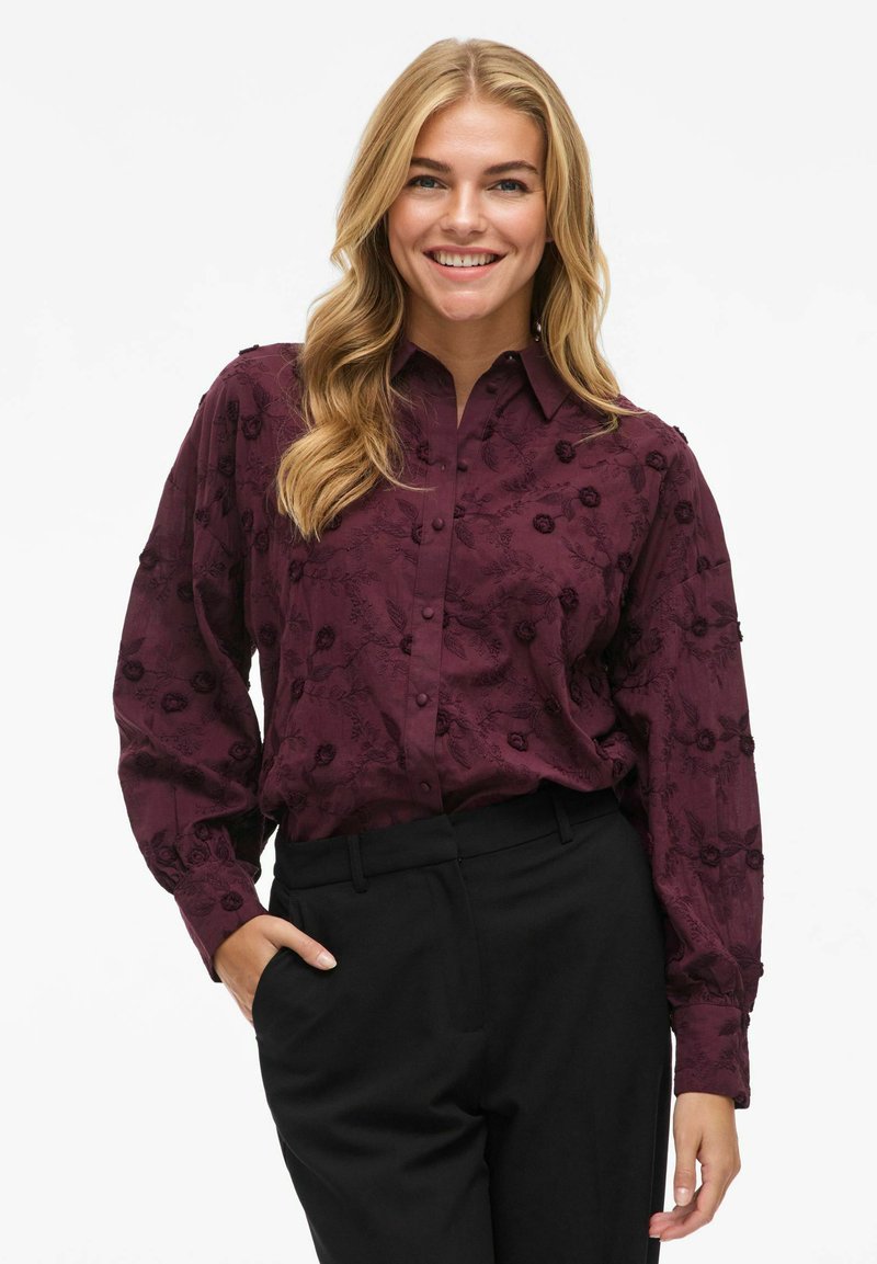 Blouse bordeaux avec broderie florale, design à boutons et manches longues ; associée à un pantalon noir pour un look contrasté.
