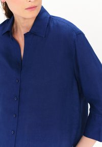 Marineblauwe linnen shirt met een knoopsluiting aan de voorkant, een kraag en driekwartmouwen; heeft een gladde textuur en een subtiele glans.