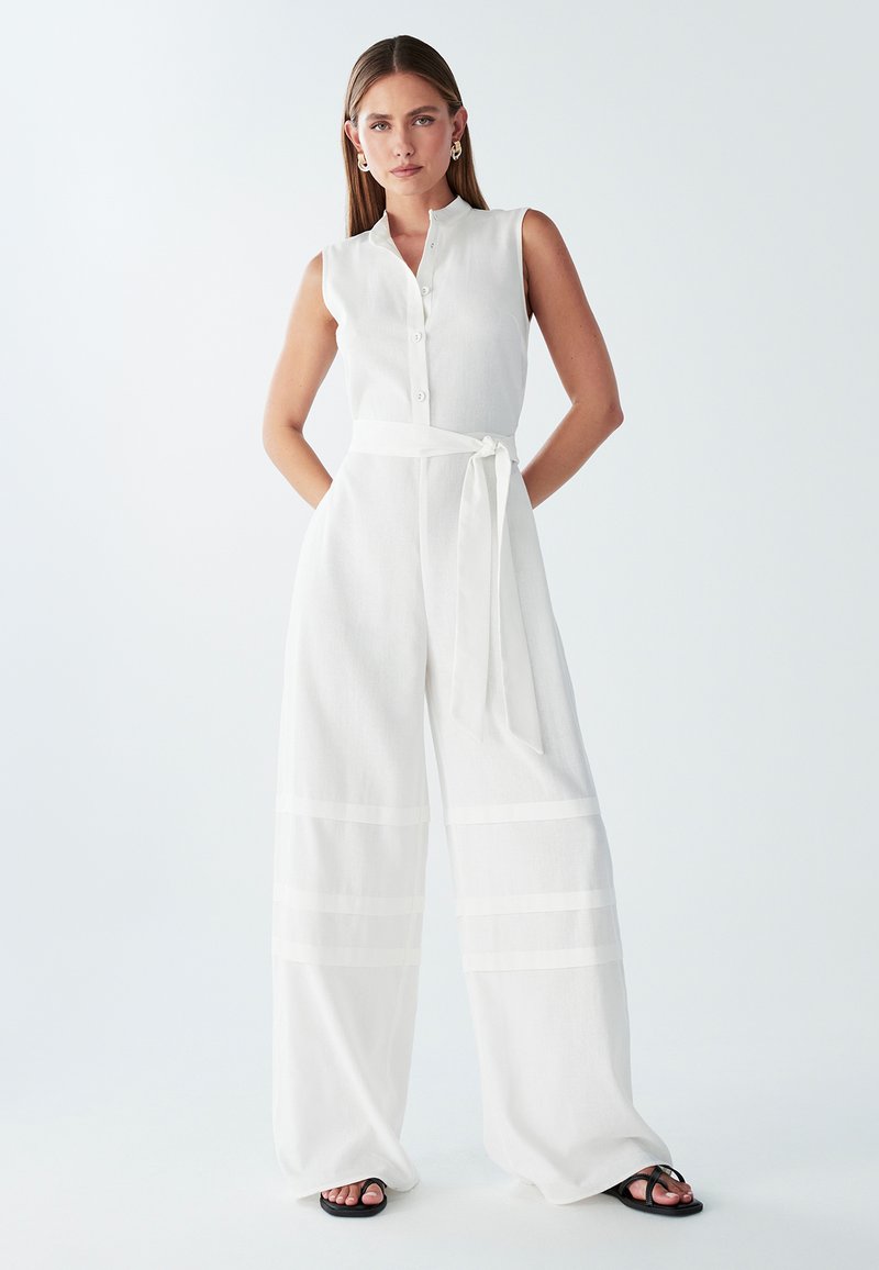Mono blanco de lino con cuello de solapa, botones frontales, piernas anchas y un cinturón de tela atado a la cintura con acentos de rayas horizontales.
