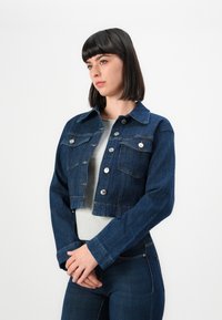 Young woman with short dark hair wearing a dark blue cropped denim jacket over a light gray shirt, standing against a white background.