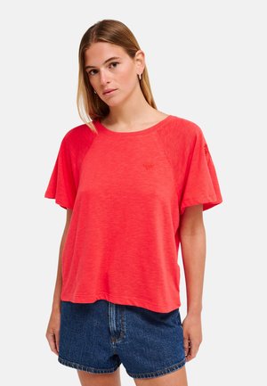 Femme portant un haut rouge ample à manches courtes et un short en jean bleu, debout devant un fond clair uni.