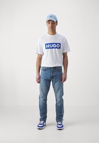 Tricou alb cu logo-ul albastru "HUGO", șapcă albastră deschisă, blugi albaștri estompați și adidași alb-albaștri, stând în fața unui fundal neutru.