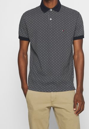 Marineblaues Poloshirt mit einem weißen Punktmuster, ausgestattet mit einem Kragen und kurzen Ärmeln. Begleitet von khakifarbenen Hosen.