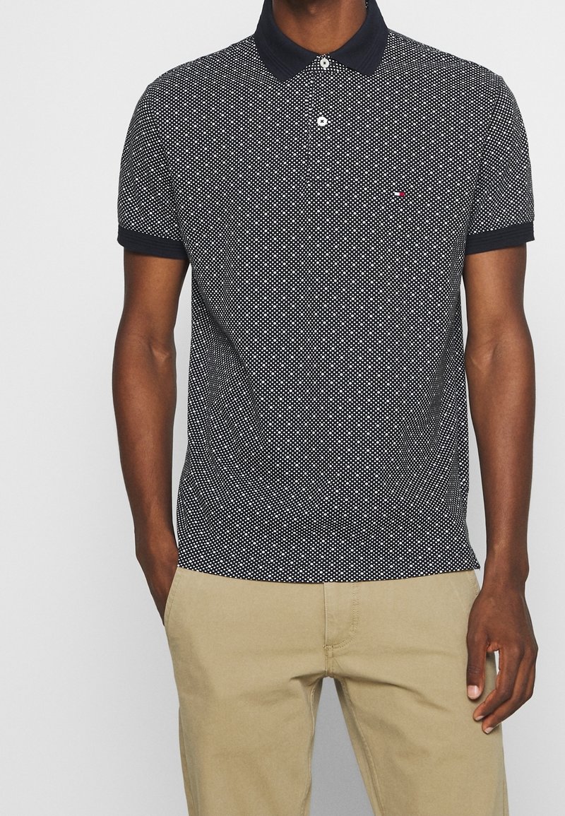 Polo shirt marine à motif de pois blancs, avec un col et des manches courtes. Accompagné de pantalons kaki.