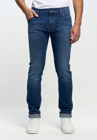 Jeans de mezclilla azul con un ajuste slim, diseño de cinco bolsillos, con un desgaste sutil y puños enrolados, combinados con zapatillas blancas.