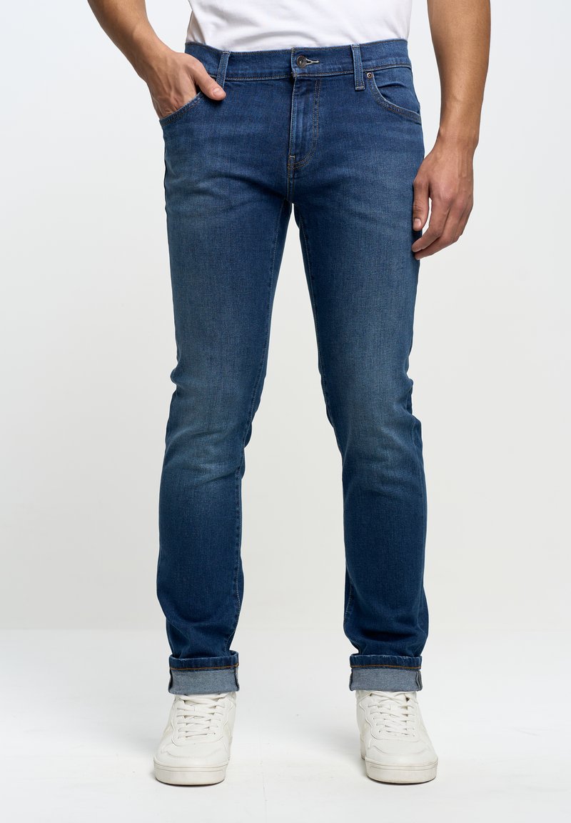 Jeans de mezclilla azul con un ajuste slim, diseño de cinco bolsillos, con un desgaste sutil y puños enrolados, combinados con zapatillas blancas.