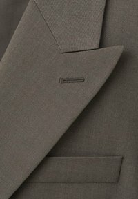 Gros plan sur le revers et la poche d'une veste marron avec boutonnière visible, montrant une texture de tissu lisse et tissé serré.