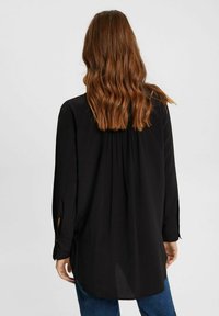 Maglietta nera a maniche lunghe con una vestibilità casual, caratterizzata da un design sulla parte posteriore con pieghe e polsini con bottoni. Abbinata a jeans blu.