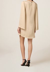 Žena v béžových šatech s dlouhým rukávem a nařasenými rameny, s černými lakovanými podpatky na klínu, na fotografii z pohledu zezadu na jednolitém pozadí.